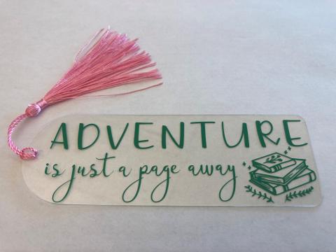 Acrylic bookmark with a tassel and the words "Adventure is just a page away" and an image of a stack of books