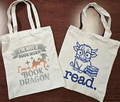 Two tote bags: One with a dragon that says "I'm not a book worm I'm a book dragon" an the other has an image of a cow that says "read"